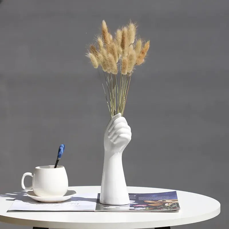 White hand-shaped vase with dried grass on round table, cup and magazine beside it