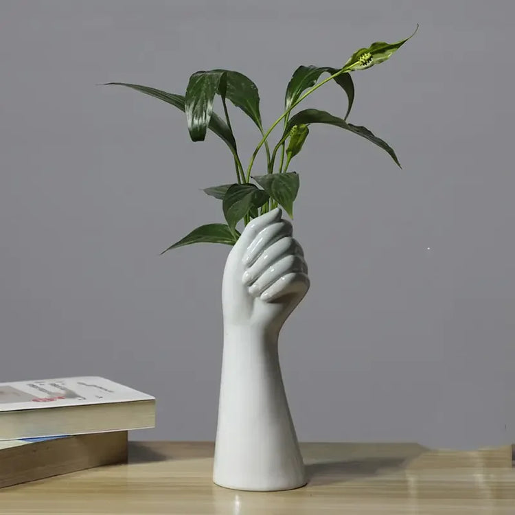 White ceramic hand vase holding green plant on wooden desk with books