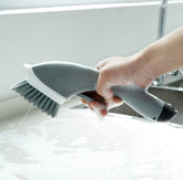 Hand using a grey dish scrub brush with soap in a kitchen sink