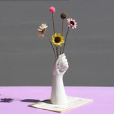 White hand-shaped vase holding colorful artificial flowers on a purple table